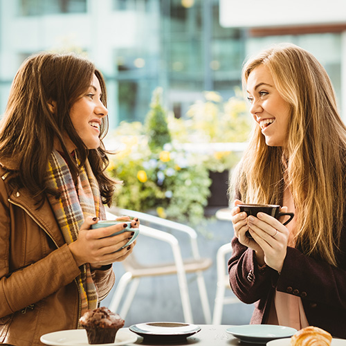 Two women talking at an outdoor cafe holding mugs of coffe